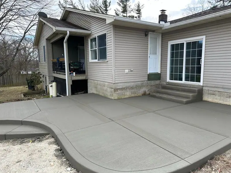 Freshly poured gray concrete patio with a curved edge and steps behind a home