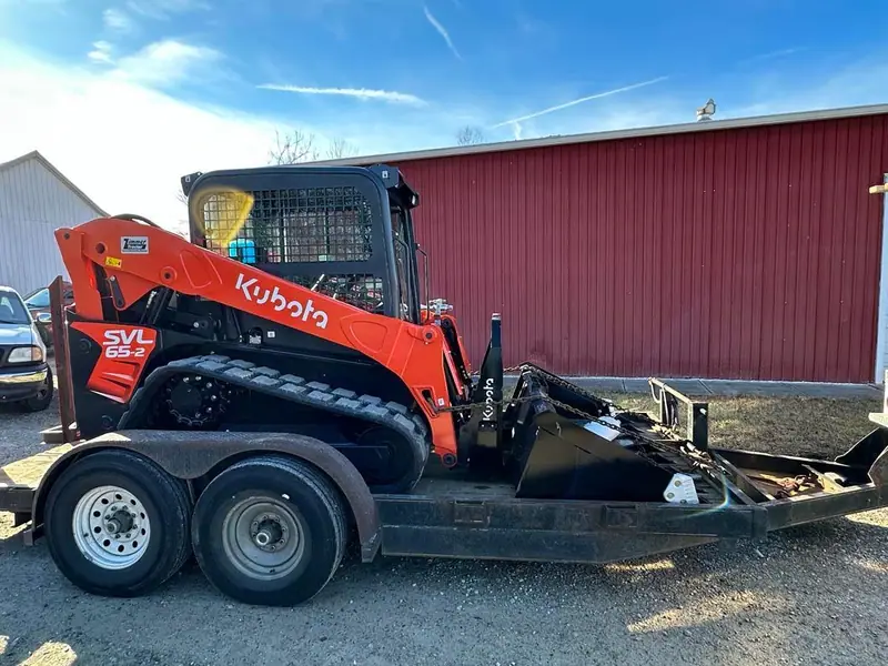Orange compact track loader on a trailer parked beside a red barn